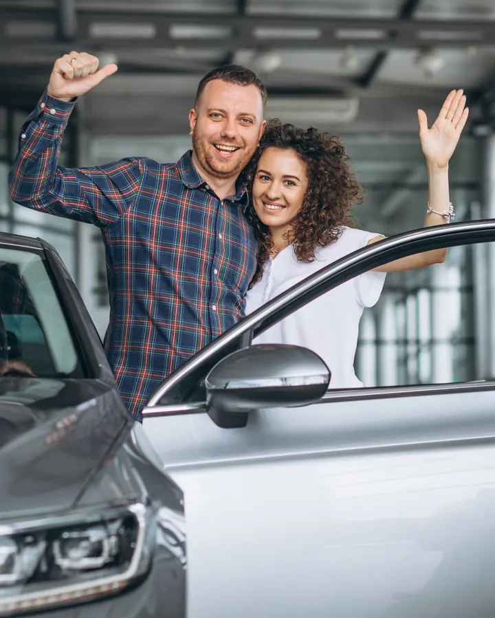 Casal comemorando ao lado do carro novo em uma concessionária